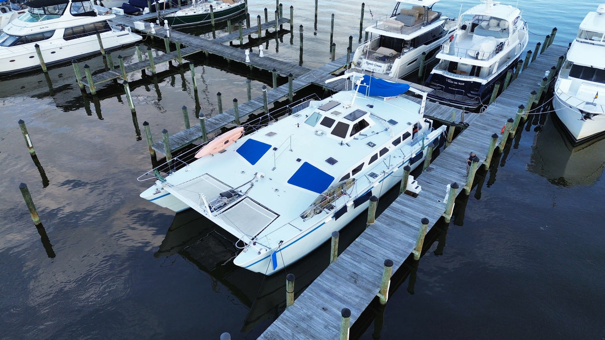 Aerial drone photo of our cruising catamaran docked at the marina, showing solar panels on the coachroof and stern arch, blue hatch covers, kayaks, and forward trampolines